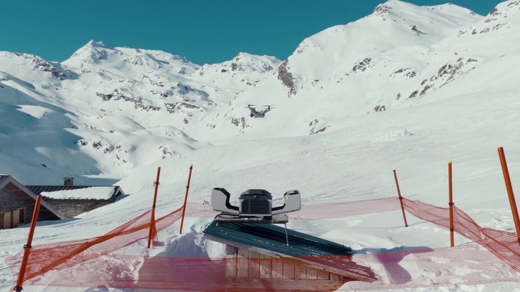 Paysage de montagne enneigée avec, au premier plan, une motoneige garée derrière des filets de sécurité orange. Des sommets enneigés et un ciel bleu clair sont visibles à l'arrière-plan.