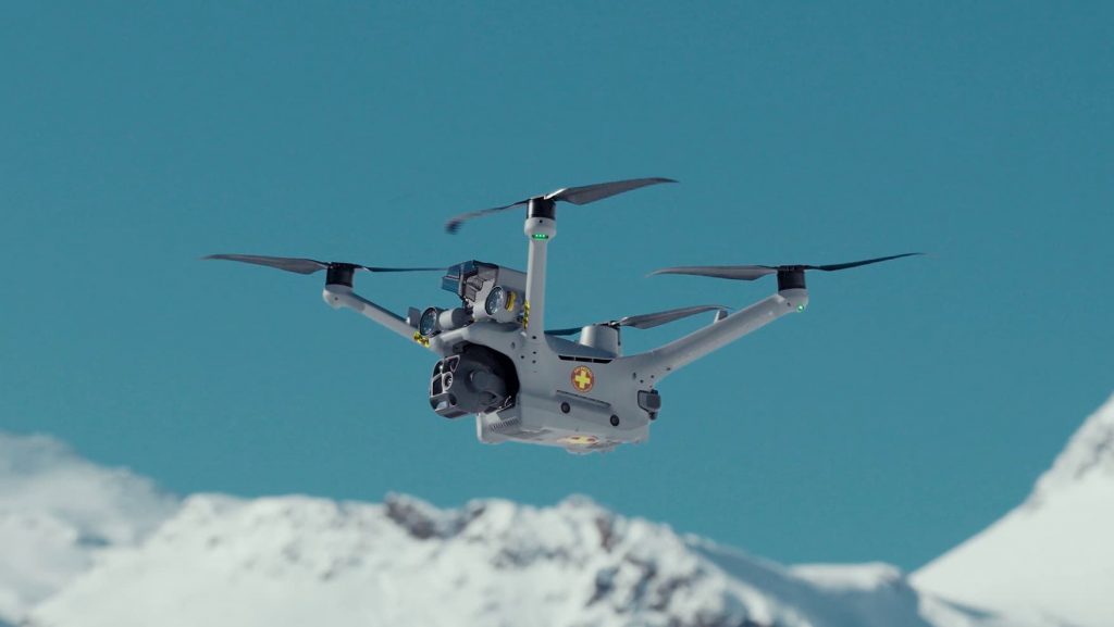 Un drone équipé de caméras et d'un symbole médical plane dans les airs au-dessus des montagnes enneigées, sous un ciel bleu limpide.