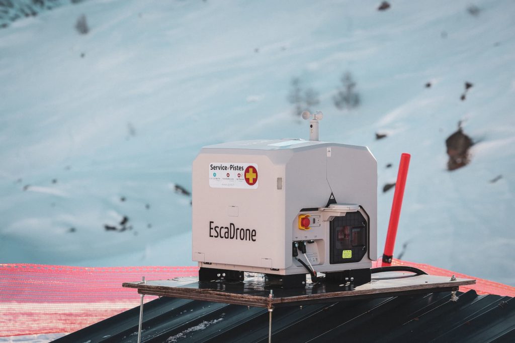 Un EscaDrone blanc est monté sur un toit métallique, avec un filet de sécurité rouge et un terrain montagneux enneigé en arrière-plan. L'appareil est équipé d'un panneau de commande et de marques de service d'urgence.