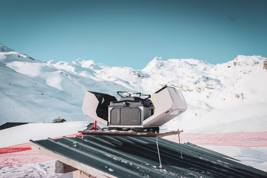 Un drone compact et moderne est stationné sur un toit métallique dans un paysage de montagne enneigé sous un ciel bleu clair, avec des pistes de ski et des montagnes visibles en arrière-plan.
