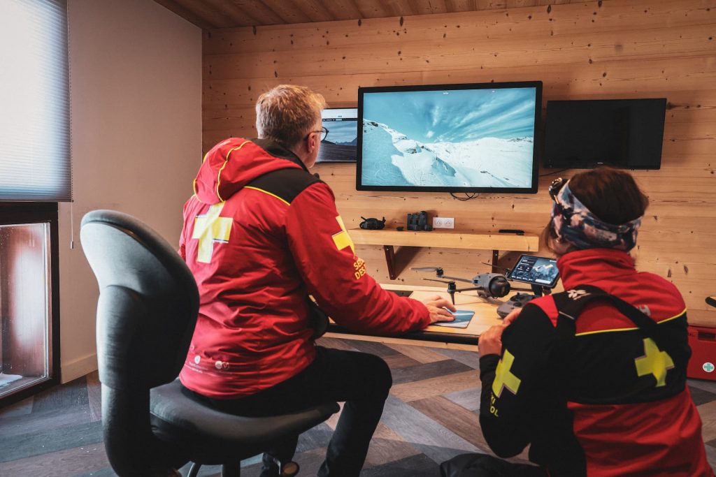 Deux personnes portant des vestes rouges à croix jaune sont assises dans une pièce en bois, surveillant une scène de montagne enneigée sur un grand écran. Elles semblent concentrées, entourées d'équipements techniques et de lumière naturelle.