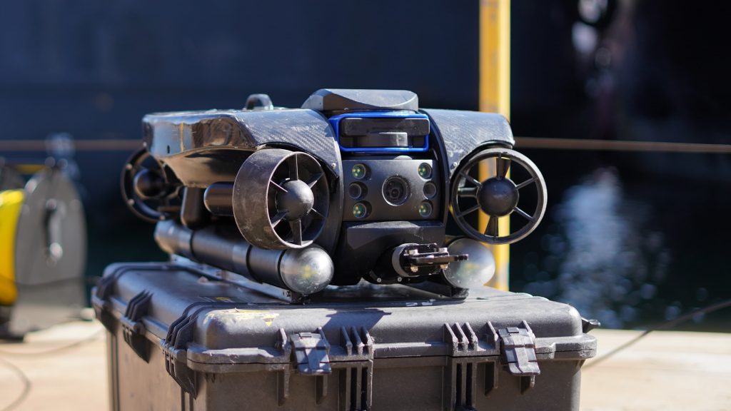 Un drone sous-marin noir équipé d'hélices et de caméras est placé sur un boîtier rigide près de l'eau, avec des équipements industriels visibles à l'arrière-plan.