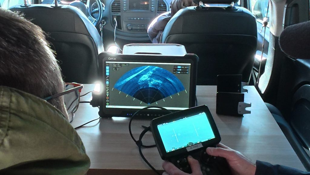 Deux personnes sont assises dans un véhicule, visionnant des images sonar sous-marines sur une tablette et contrôlant l'équipement à l'aide d'un appareil portatif placé sur une table entre les sièges avant.