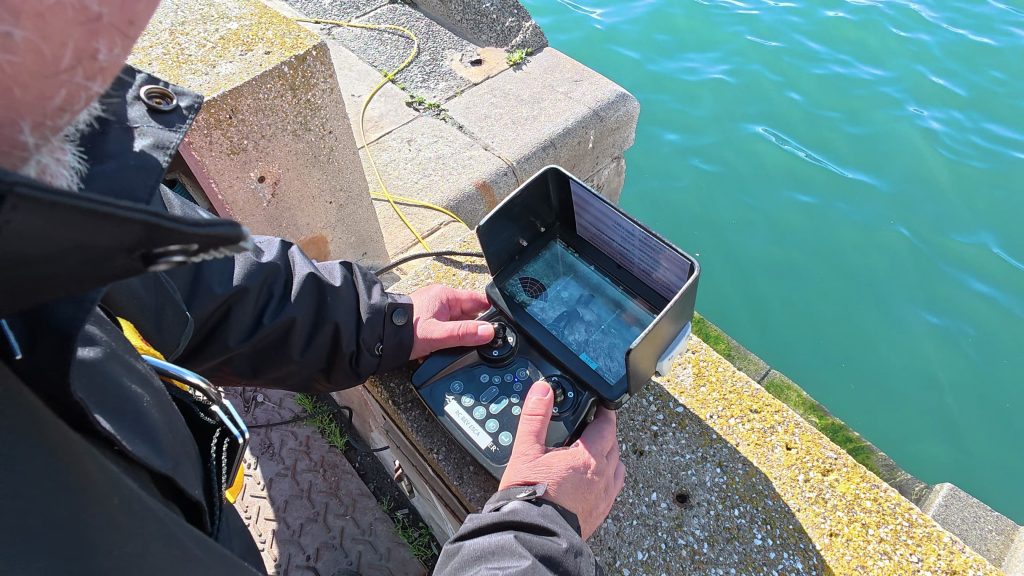 Une personne actionne une télécommande avec écran, peut-être pour un drone ou un véhicule sous-marin, alors qu'elle se tient sur un quai en béton au bord de l'eau bleue.