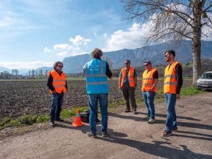 Cinq personnes portant des gilets de sécurité orange se tiennent sur un chemin de terre près d'un champ, écoutant une personne vêtue d'un gilet bleu. On aperçoit des montagnes et un arbre en arrière-plan ; un cône orange et une voiture garée se trouvent à proximité.