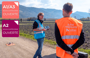 Deux personnes se tiennent sur un chemin de terre près d'un champ ; l'une d'elles porte un gilet orange portant l'inscription « ÉLÈVE TÉLÉPILOTE », indiquant sa participation au programme de formation « Formation télépilote de drone professionnel Parcours Open », tandis qu'un drone repose au sol. Des panneaux indiquant les sous-catégories A1/A3 et A2 sont visibles à gauche.