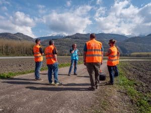 Cinq personnes, dont quatre portent des gilets de sécurité orange, se tiennent sur un chemin de terre à la campagne et discutent. En arrière-plan, on aperçoit des montagnes, des champs et un ciel partiellement nuageux. L'une d'entre elles tient un bloc-notes.