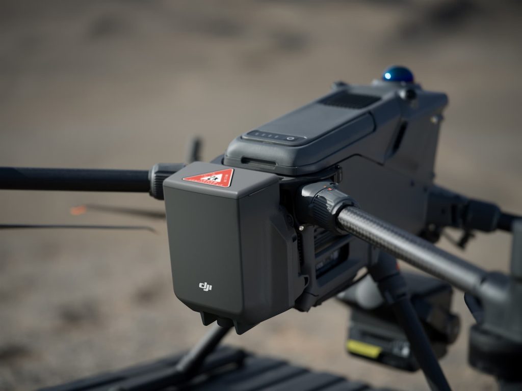 Gros plan sur le Parachute DJI AP100 pour Matrice 400 (C5/C6), montrant son étiquette d'avertissement, sa caméra et son module de capteurs à l'extérieur, sur fond de paysage flou.