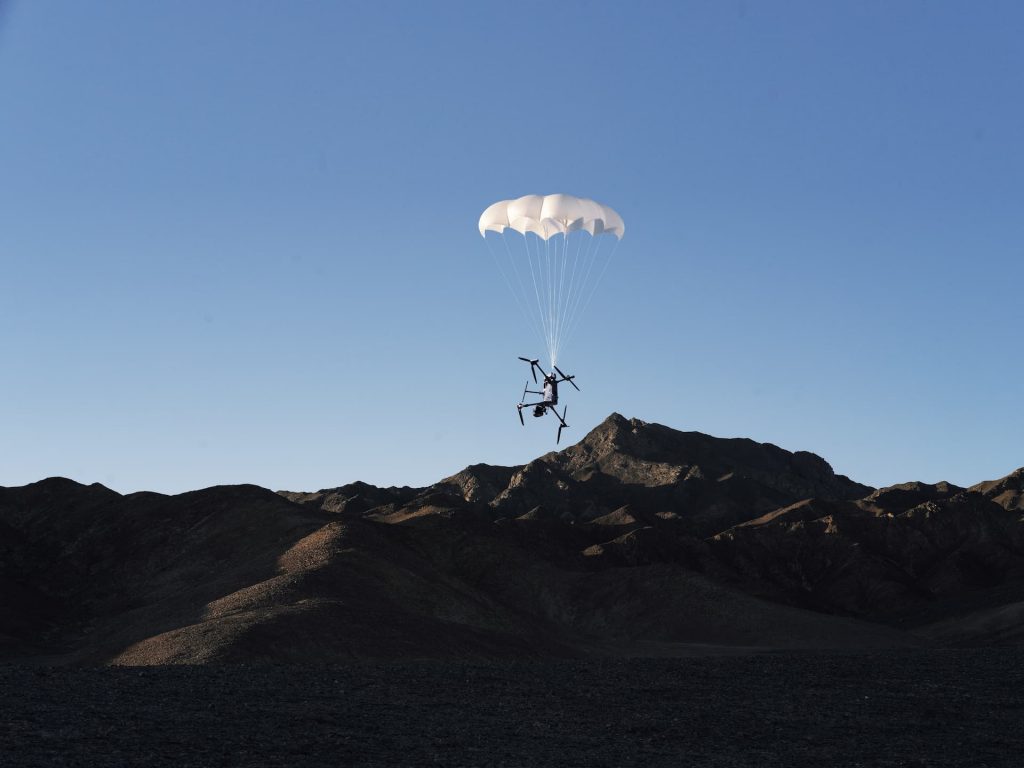 Le parachute DJI AP100 pour Matrice 400 (C5/C6) se déploie alors que le drone descend dans un ciel bleu limpide, avec en arrière-plan la silhouette de montagnes escarpées et d'un relief rocheux.