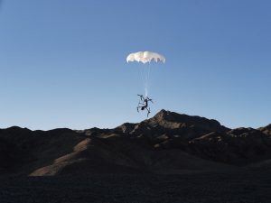 Le parachute DJI AP100 pour Matrice 400 (C5/C6) se déploie alors que le drone descend dans un ciel bleu limpide, avec en arrière-plan la silhouette de montagnes escarpées et d'un relief rocheux.