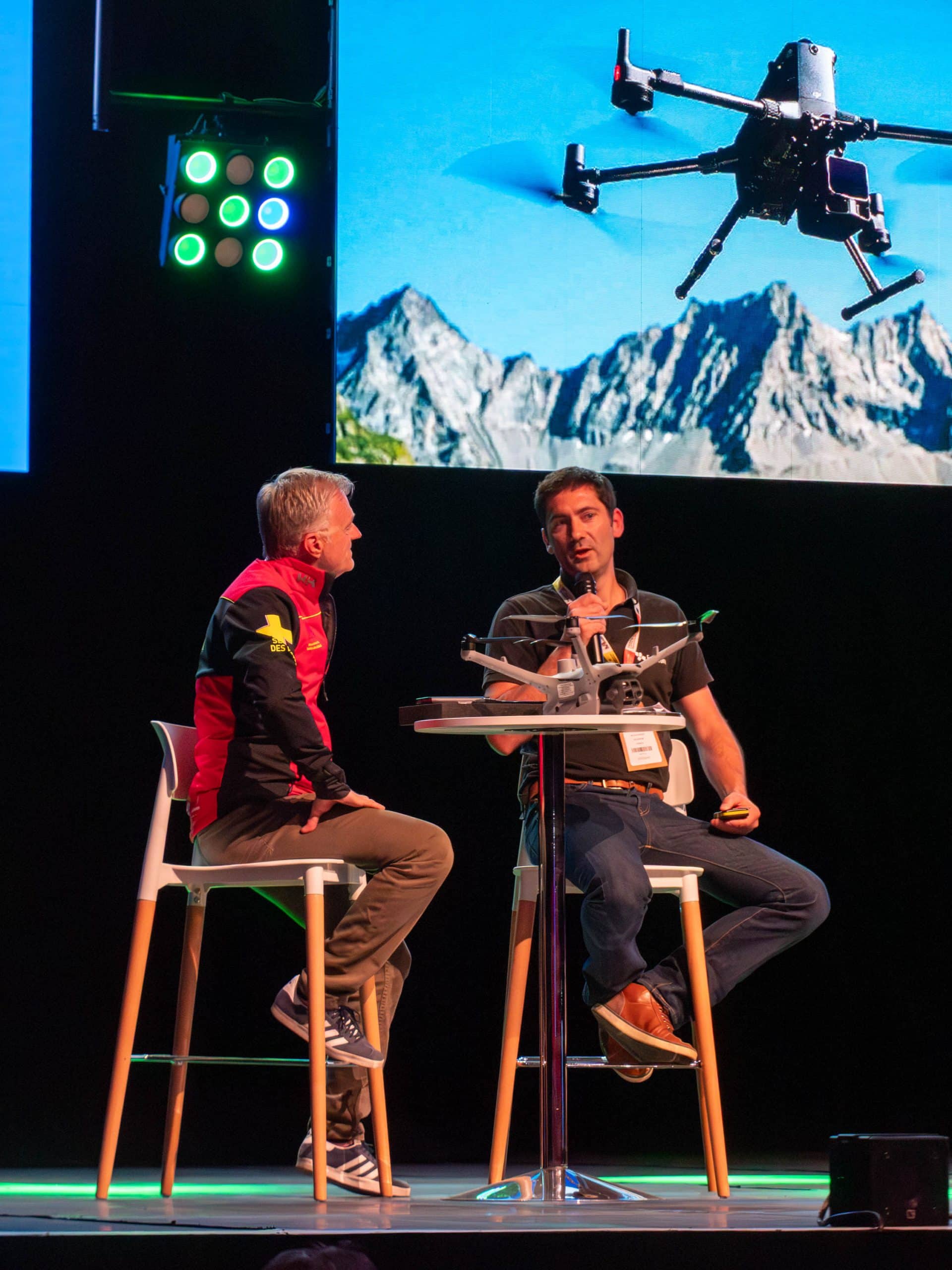 Deux hommes sont assis sur des chaises hautes autour d'une petite table sur scène et discutent d'un modèle de drone posé sur la table. Un écran derrière eux montre un drone volant près de montagnes. On aperçoit les projecteurs de la scène et les badges de l'événement.
