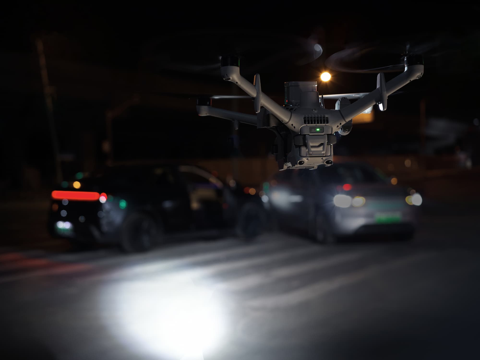 Un drone DJI Dock vole en stationnaire de nuit, projetant un faisceau lumineux sur une route où l'on distingue deux voitures floues en arrière-plan.