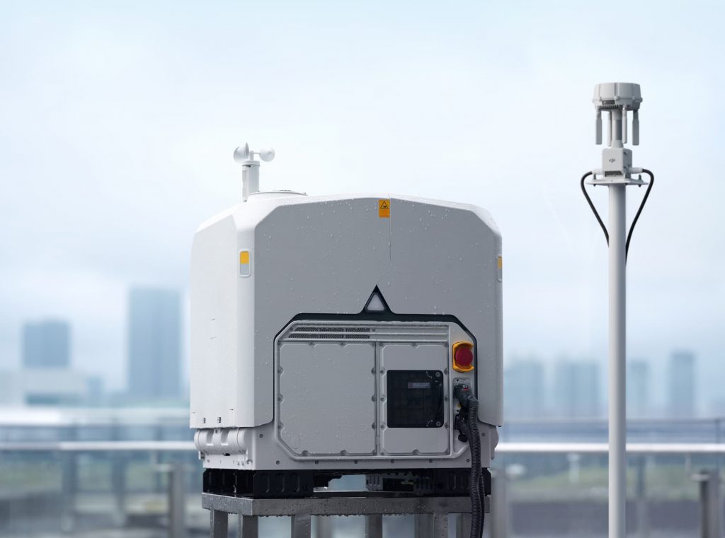 Une station d'accueil pour drone DJI Dock 4, blanche et de forme cubique, installée à l'extérieur, avec un capteur météo sur le dessus et un mât d'antenne séparé à proximité.