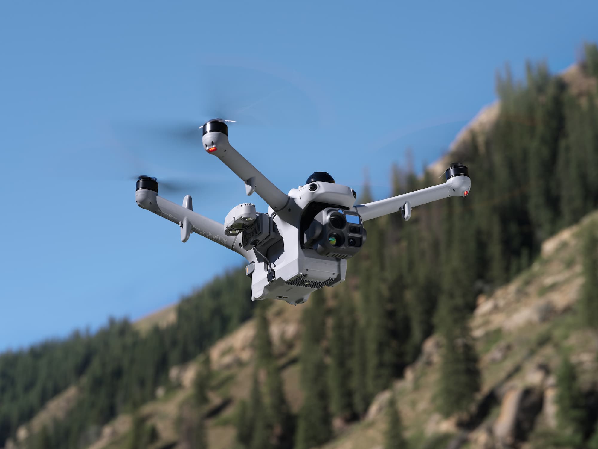 Un drone quadricoptère gris équipé d'un projecteur DJI AL2D pour Matrice 5D et 5TD, volant devant un décor de montagnes et d'arbres.
