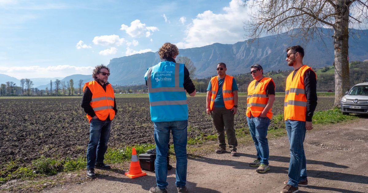 Cinq personnes portant des gilets de sécurité orange se tiennent sur un chemin de terre près d'un champ, écoutant une personne vêtue d'un gilet bleu. On aperçoit des montagnes et un arbre en arrière-plan ; un cône orange et une voiture garée se trouvent à proximité.