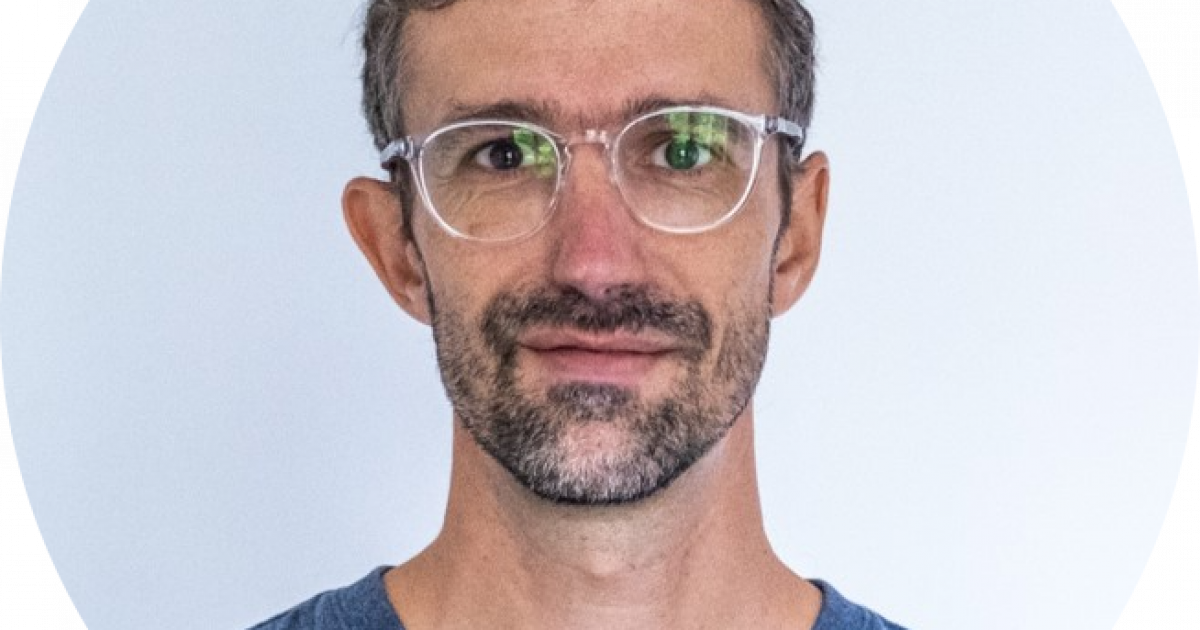 Un homme aux cheveux bruns courts et bouclés, portant une barbe et une moustache, avec des lunettes transparentes et un t-shirt bleu à col en V, debout devant un fond clair et uni, regardant directement l'appareil photo.