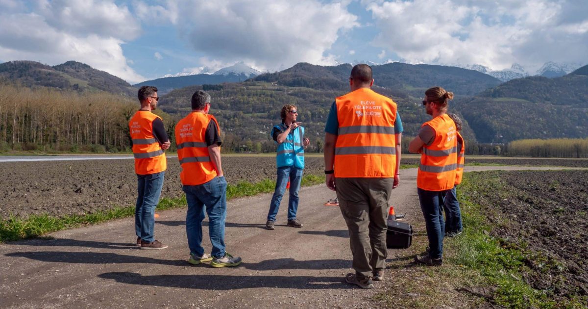 Cinq personnes, dont quatre portent des gilets de sécurité orange, se tiennent sur un chemin de terre à la campagne et discutent. En arrière-plan, on aperçoit des montagnes, des champs et un ciel partiellement nuageux. L'une d'entre elles tient un bloc-notes.