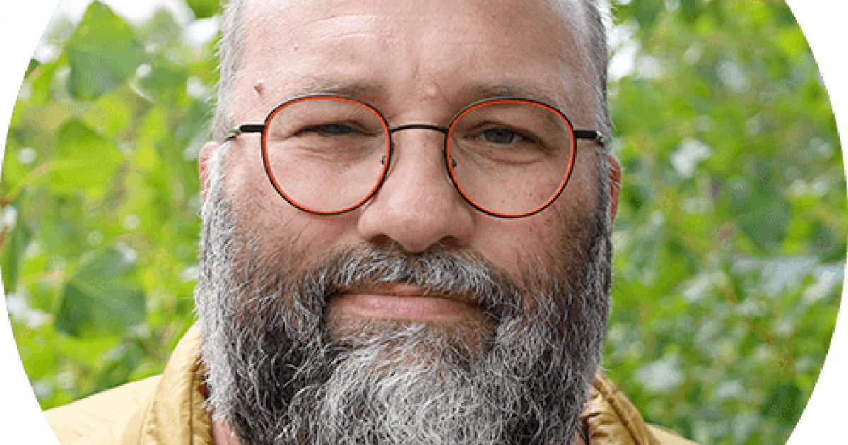 Un homme d'âge moyen, à la barbe et à la moustache grises, portant des lunettes rondes et une veste jaune, se tient à l'extérieur, devant des arbres verts et feuillus.