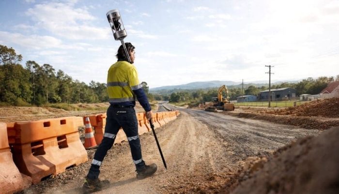 Un ouvrier de la construction en tenue de sécurité marche le long d'un chemin de terre sur un chantier de construction, transportant un équipement d'arpentage avec un LiDAR Hovermap GX1. Des barrières orange bordent la route tandis qu'une pelleteuse travaille à l'arrière-plan sous un ciel partiellement nuageux.