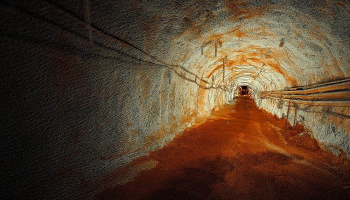 Un tunnel souterrain faiblement éclairé avec des murs rugueux et rocheux de couleur orange rouille et grise, des tuyaux et un scanner LiDAR Hovermap GX1 monté sur le mur de gauche, le tout menant dans l'obscurité au loin.