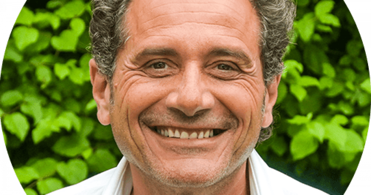 Un homme d'âge moyen aux cheveux noirs bouclés et au large sourire porte une chemise blanche et se tient en plein air devant un feuillage vert.