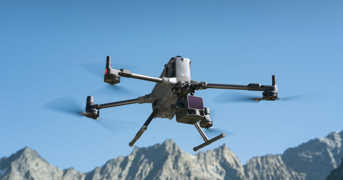 Un drone quadcopter DJI L3 LiDAR équipé d'une caméra vole devant des montagnes escarpées et ensoleillées sous un ciel bleu clair.