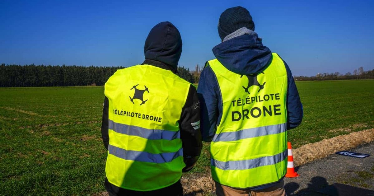 Deux personnes portant des gilets jaunes portant l'inscription « Telepilote Drone » se tiennent au bord d'une route, près d'un champ herbeux, avec en arrière-plan un ciel bleu éclatant et des arbres. Cette scène met en évidence la présence de télépilotes certifiés pilotant des drones dans un cadre naturel et à ciel ouvert.