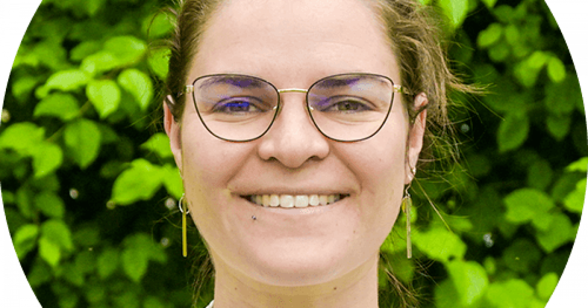 Une personne à la peau claire, portant des lunettes et une chemise blanche, sourit devant un fond de plantes à feuilles vertes.