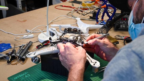 Dans un centre de réparation animé, une personne portant un masque facial répare un drone, travaillant sur son circuit imprimé exposé au milieu d'un établi encombré de fils, d'outils et d'équipements électroniques.