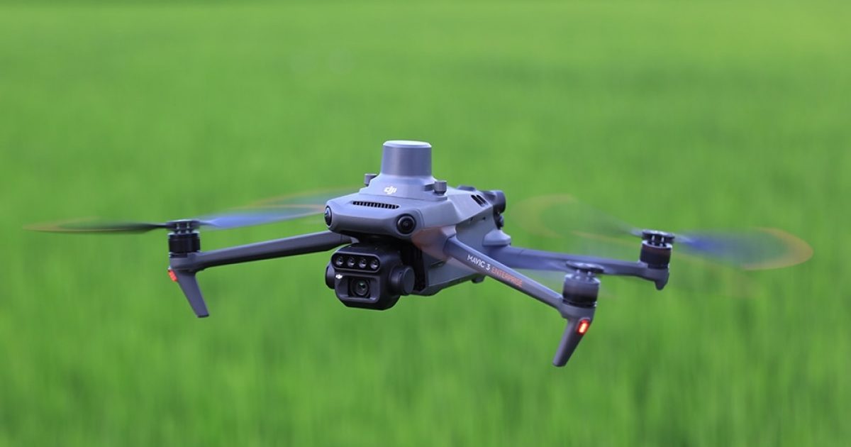 Le DJI Mavic 3 Multispectral, un drone gris équipé d'une caméra et de capteurs, plane au-dessus d'un champ vert flou, avec des hélices qui tournent et de petites lumières orange qui brillent sur ses bras.